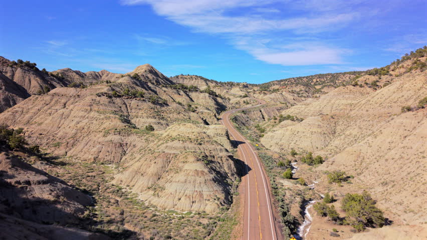 Aerial drone footage captures winding scenic highway cutting through dramatic Utah badlands with layered sedimentary rock formations, eroded hills in golden and tan hues, sparse desert vegetation.