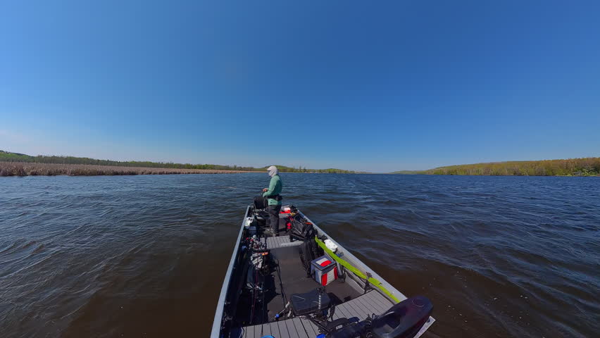 Fisherman is fishing on lake from boat casting spinning rod at daylight. Angler is standing in an equipped boat and catching fish while trolling. Man holds spinning rod while offshore during sunny day