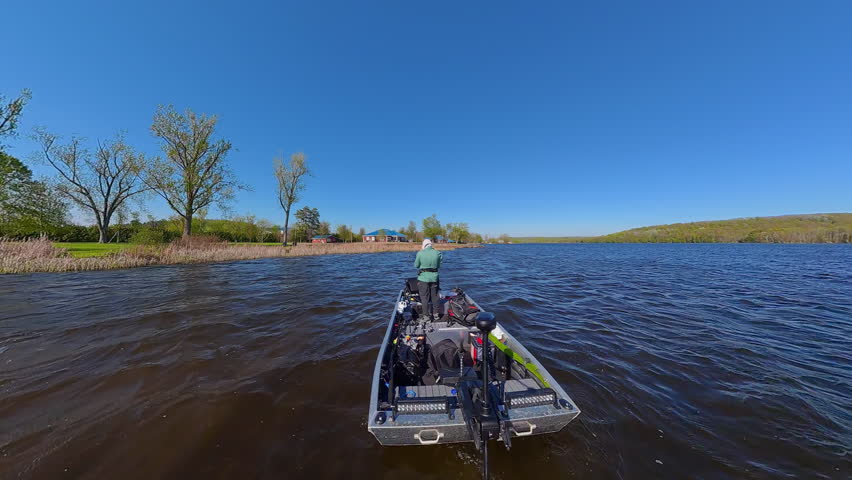 Fisherman is fishing on lake from boat casting spinning rod at daylight. Angler is standing in an equipped boat and catching fish while trolling. Man holds spinning rod while offshore during sunny day