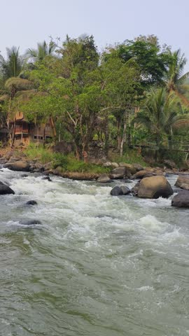 A scenic view of Citarik River flowing through a tropical landscape with large natural rocks, lush greenery, coconut palm trees, and a clear blue sky in Sukabumi, West Java, Indonesia.