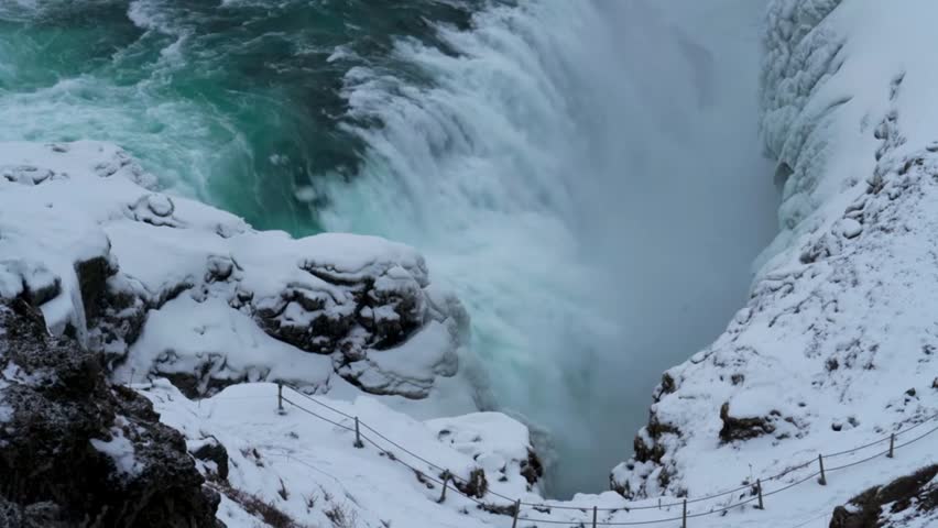 High-angle shot of powerful waterfall rushing through snowy, ice-covered rock canyon with turquoise water flowing fiercely into the frozen gorge during cold winter season.