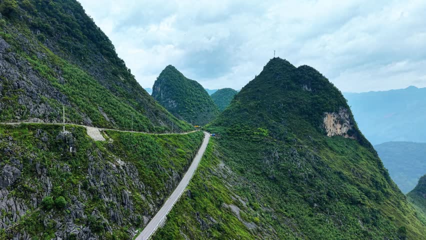 Aerial drone footage following a rugged limestone mountainside toward a dramatic hairpin bend along a mountain road in the Dong Van Karst Plateau near Meo Vac, northern Vietnam. The road wraps tightly around the steep cliff face, revealing the extreme terrain and engineering required to connect communities across the mountainous landscape. Towering rock formations dominate the frame while the winding roadway creates a striking visual contrast against the natural limestone cliffs. The forward-moving aerial perspective conveys a strong sense of adventure, elevation and exploration, making the footage suitable for Ha Giang Loop travel campaigns, motorcycle and road trip advertising, tourism promotions, destination marketing, transportation documentaries, geography content, adventure travel productions and cinematic establishing shots showcasing the spectacular mountain roads of northern Vietnam.