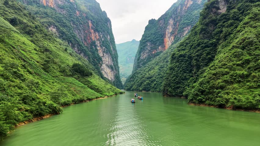 Low-altitude aerial drone footage passing close to sightseeing boats traveling into Tu San Canyon along the emerald-green Nho Que River in the Meo Vac region of northern Vietnam. The small tourist boats move between towering limestone cliffs rising dramatically on both sides of the narrow river, creating an immersive perspective of the canyon and its river-based tourism experience. The close aerial viewpoint emphasizes the scale of the vertical rock walls while capturing the peaceful movement of boats across the striking green water. The footage is suitable for Vietnam tourism advertising, Nho Que River boat tour promotions, destination marketing, adventure travel campaigns, travel documentaries, social media content, nature productions and cinematic travel sequences showcasing Tu San Canyon.