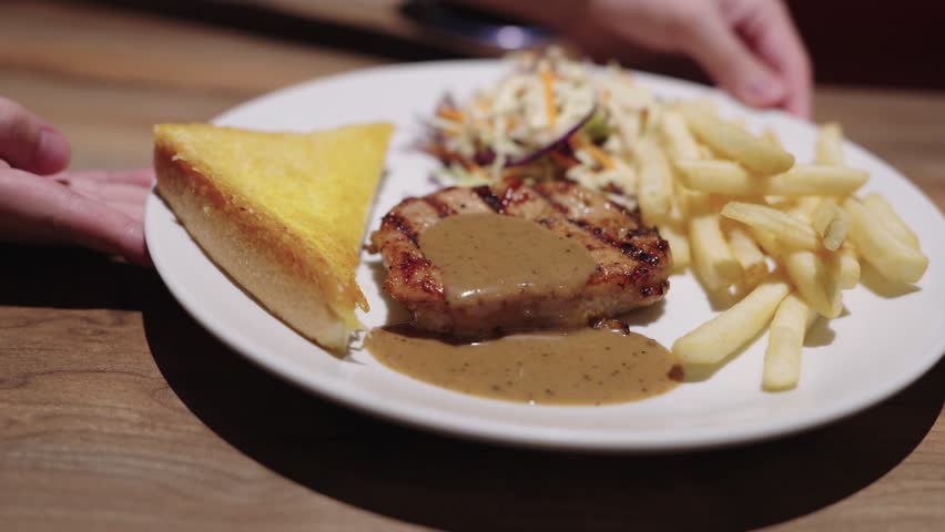 Grilled pork steak with gravy sauce served on a white plate with french fries, coleslaw salad, and toasted bread. A person holding the plate in a restaurant setting with warm lighting.