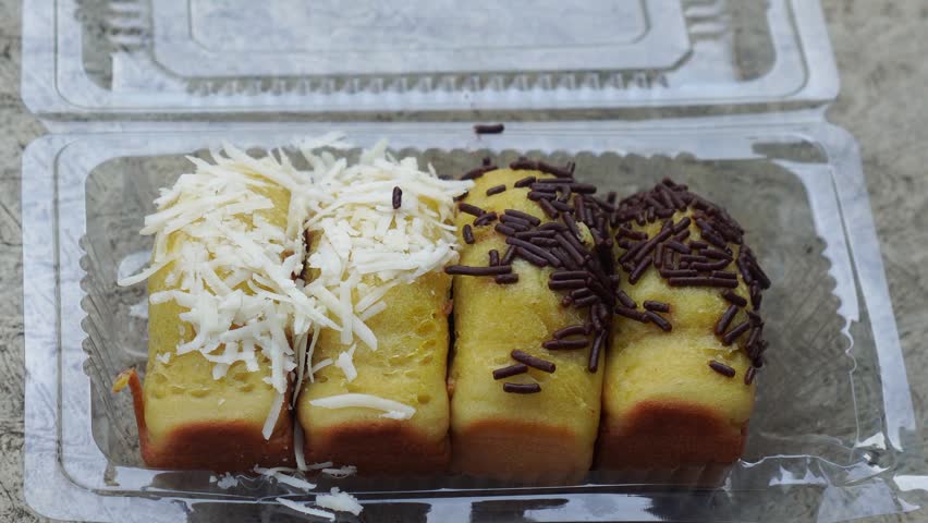 traditional Indonesian sweet sponge cakes topped with grated cheese and chocolate sprinkles in a clear plastic container, resting on a raw concrete surface. 