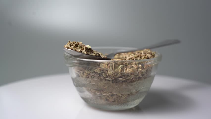 Close-Up Shot of Anise Seeds in a Glass Bowl with a Spoon, Rotating on a White Surface against a Gray Background