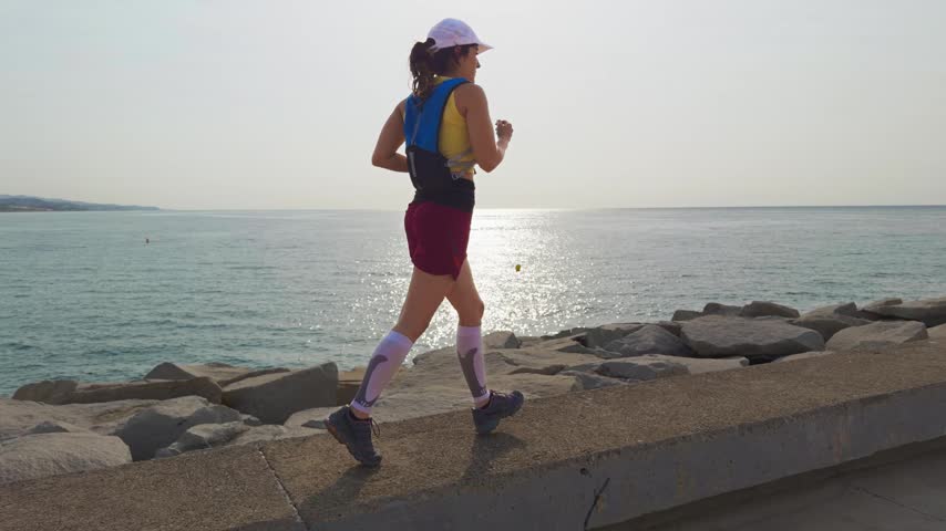 Woman runs steadily along a coastal path. She wears a white cap and athletic gear. The sea glows under the setting sun. Her expression shows focus and energy