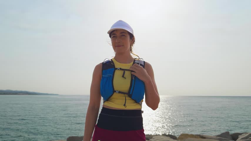Woman stands on beach. Wears yellow shirt. Blue life vest. White helmet. Smiling. Man