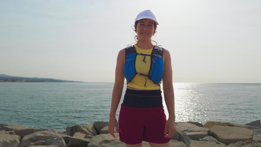 Woman smiles by the sea. She wears a life vest and white cap. Man
