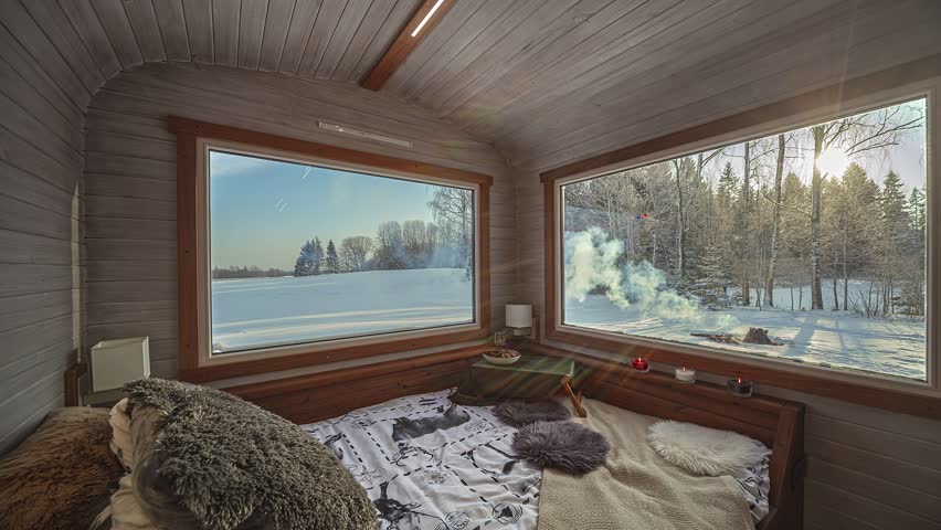 Warm interior of a rustic cabin room, large windows reveal a peaceful winter forest and field at sunset. Snow covers the ground.