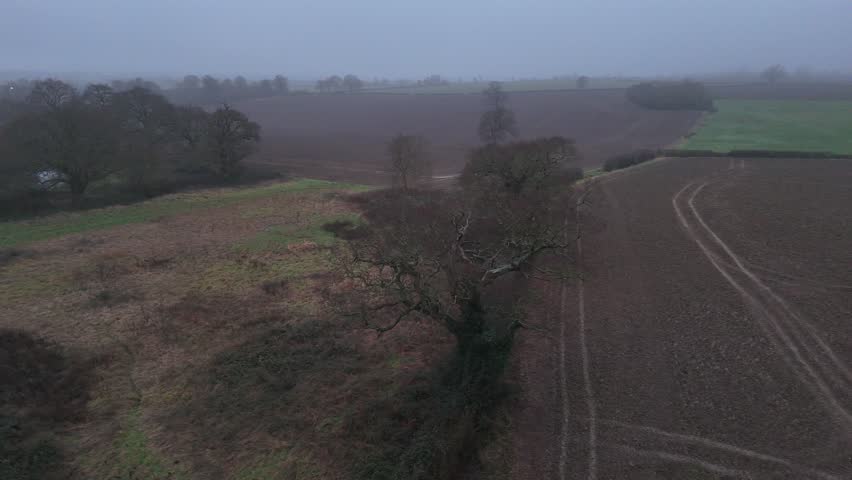 Aerial view features vast agricultural fields during gloomy winter afternoon with heavy mist and fog settling over dormant trees and countryside landscape in England cinematic footage.