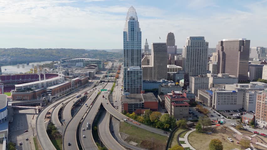 High angle aerial footage of downtown Cincinnati Ohio featuring Great American Ball Park and busy highway interchange with traffic flow under clear blue sky during autumn season.