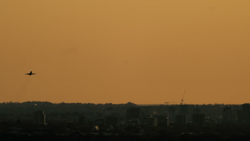 Airplane climbs into the sky above the dark buildings of Perth at sunset with golden light filling the horizon during evening
