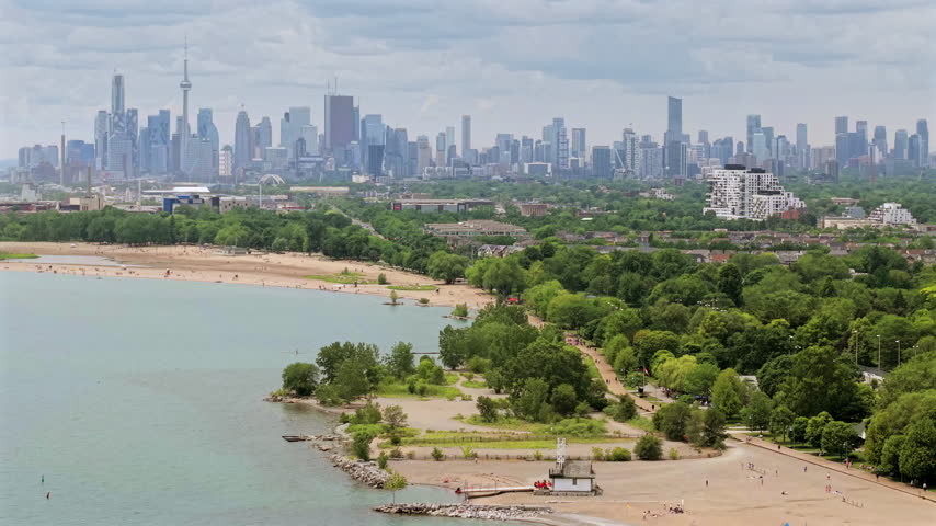 Zoomed panning aerial drone video over the eastern Toronto Beaches community with shoreline, piers, lifeguard station and downtown skyline in the distance.