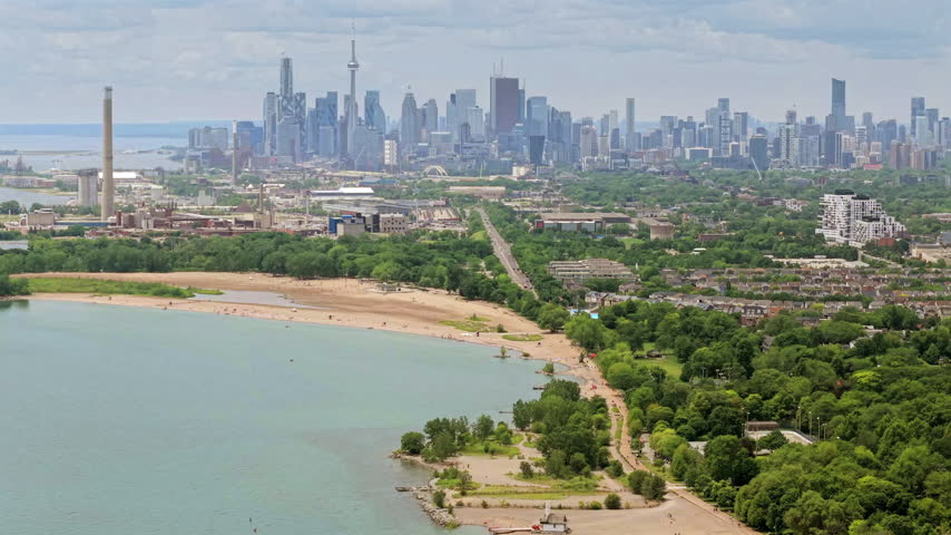 Zoomed panning aerial drone video over the eastern Toronto Beaches community with shoreline, piers, lifeguard station and downtown skyline in the distance.