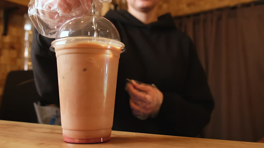 Barwoman hand stirs iced coffee with cherry juice in plastic cup closeup. Barista in gloves prepares tasty cocktail for guest in coffeeshop