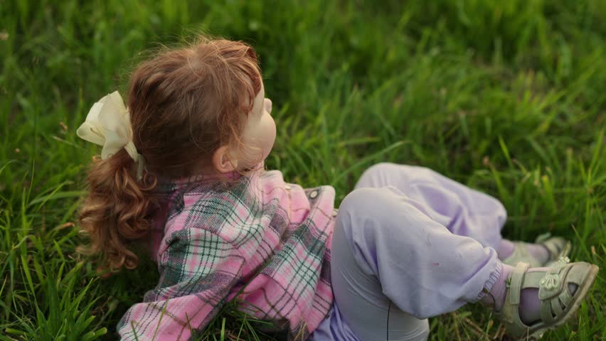 Little girl with red hair lying in green grass and sticking out her tongue. Happy child enjoying nature in summer park. Outdoor leisure, childhood fun concept.
