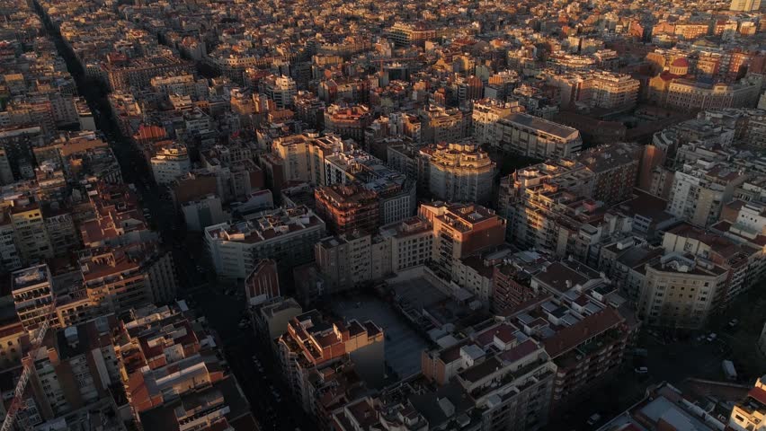 Aerial view of dense residential city block in Barcelona Spain featuring urban architecture with golden sunset sunlight casting long shadows across rooftops during peaceful morning scene.