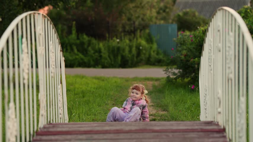 Little girl playing on grass near decorative garden bridge. Toddler exploring nature outdoors during summer day. Child walking and crouching in rural park land.