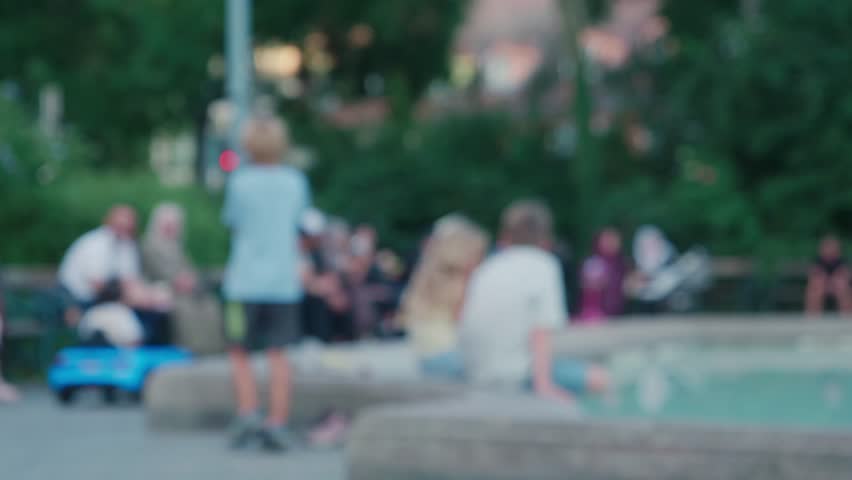 Blurred out of focus background of people relaxing and sitting near fountain in public park during summer.