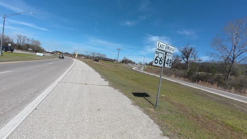 First-person POV footage of riding a bicycle along Historic Route 66 in Oklahoma. The clip passes a visible Route 66 highway sign before continuing along a wide paved shoulder and through a rural intersection with road signs, passing traffic, utility poles, and roadside scenery. No audio.