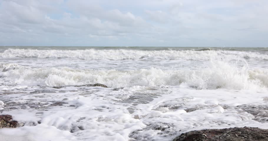 Ocean waves crashing onto a rocky shoreline with white sea foam under a cloudy blue sky, natural coastal seascape footage.