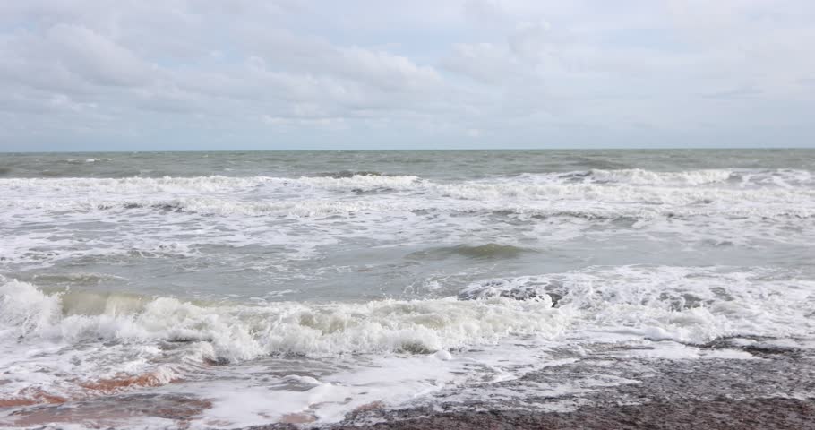Ocean waves crashing onto a rocky shoreline with white sea foam under a cloudy blue sky, natural coastal seascape footage.