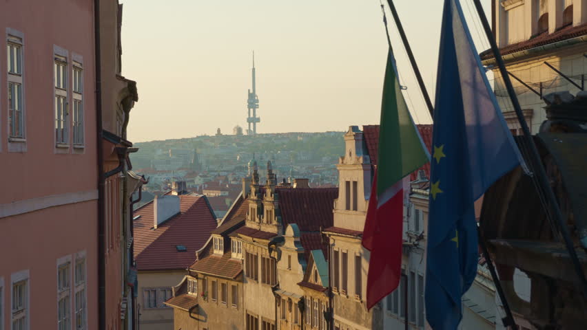 European Union flag hangs on a historic Prague street with traditional architecture. The city skyline features a famous television tower in morning light. Ideal for Czech travel and tourism.