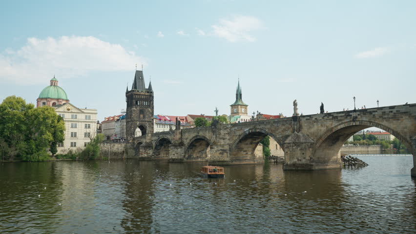 Historic Charles Bridge stands over Vltava River in Prague. A small tourist excursion boat sails under stone arches past city landmarks. Ideal for European vacation and tourism travel design.