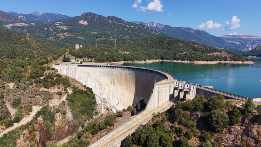 Aerial view of Baells Dam curving across a rocky gorge, holding a turquoise reservoir in Catalonia, Spain, with forested mountains, cliff bands, and a ruined hilltop castle in the distance