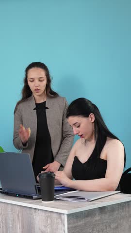 Two businesswomen working at an office desk with a laptop, documents, and a blue folder while discussing tasks against a blue background.