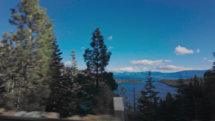 View of Emerald Bay at Lake Tahoe with island and mountains, partially obscured by blurred trees while driving along a scenic road in California