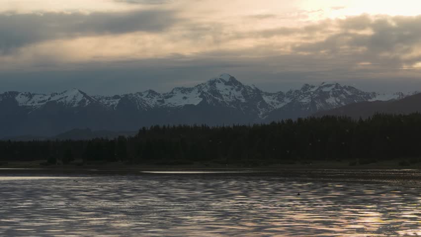 Panoramic view of a rugged mountain range with snow-covered peaks overlooking a calm lake at dusk.