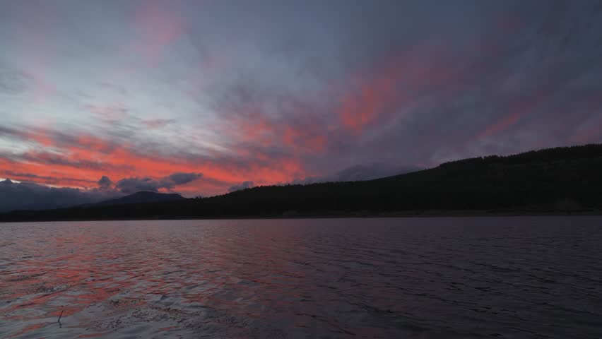 Dramatic sunset view over a tranquil lake, featuring vibrant red and purple clouds reflecting on the water, with silhouetted mountain ridges and forest in the distance.