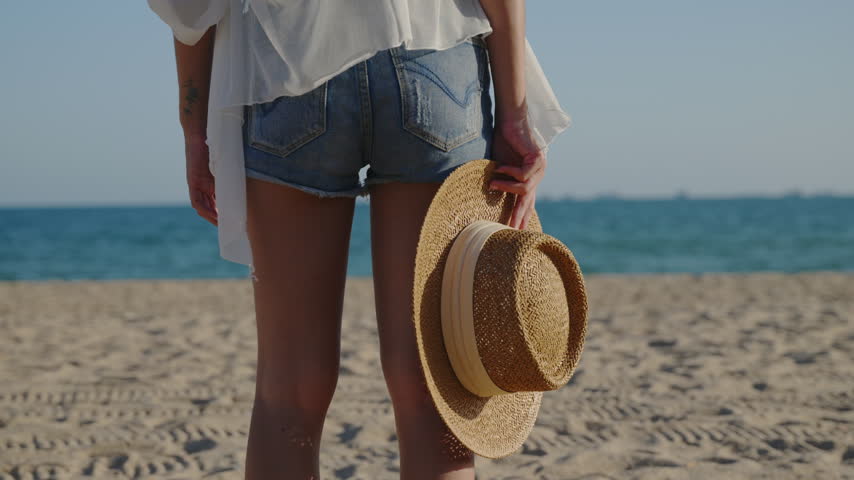 Rear view of woman, standing, hold hat, beach travel concept during sunny summer day, traveler wearing denim shorts with selective focus on straw hat and blue ocean background.