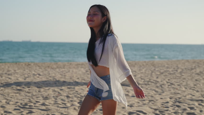 Young woman on beach during summer seeks to relax and enjoy lifestyle, standing with outstretched arms on sandy shore under sunny sky while feeling free on weekend travel vacation.