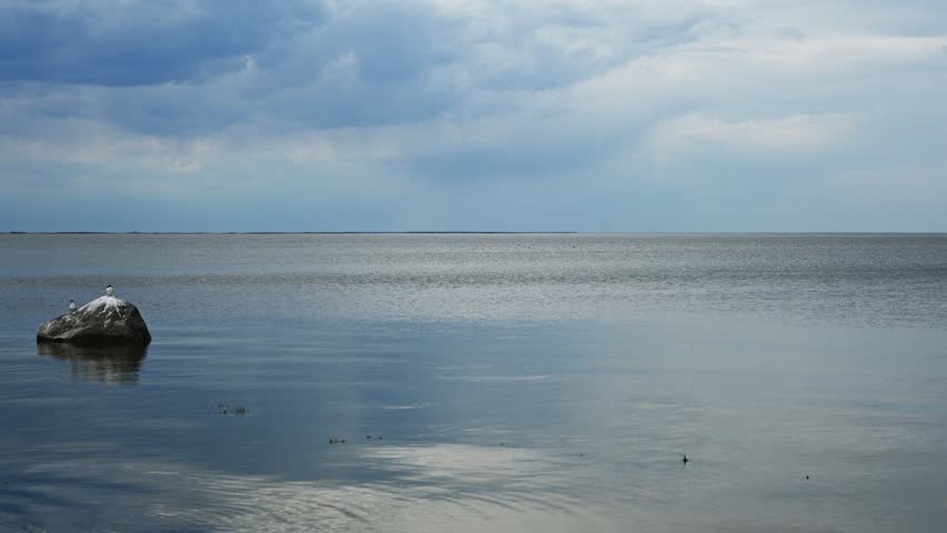 View across a large calm lake with terns on a rock and a sky full of blueish clouds