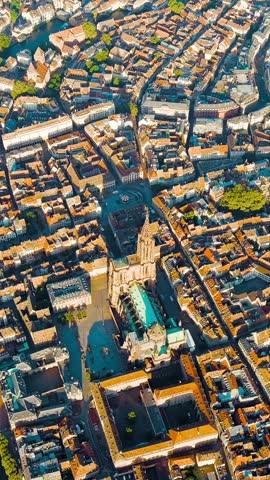 Vertical video. Strasbourg, France. Notre-Dame de Strasbourg dominates the historic city center from above, its distinctive single spire reaching 142 meters and making it the tallest medieval structure ever built. Constructed between 1176 and 1439, the edifice showcases Rayonnant Gothic architecture with its asymmetrical silhouette, the planned south tower never completed due to funding constraints. The reddish-brown Vosges sandstone gives the structure its characteristic warm tone, visible across the dense network of historic streets and rooftops that surround the central square. The aerial perspective reveals its commanding position at the heart of the UNESCO-listed Grande Ile, with the spire visible from over 30 kilometers away on clear days. Bright summer morning light illuminates the urban fabric, highlighting the contrast between the ornate Gothic mass and the tightly packed historic buildings of Alsace. Aerial View, Point of interest. Rich colors
