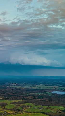 Storm clouds move at dusk with visible rain shaft falling over distant farmland and small lake in vertical aerial timelapse Latvia