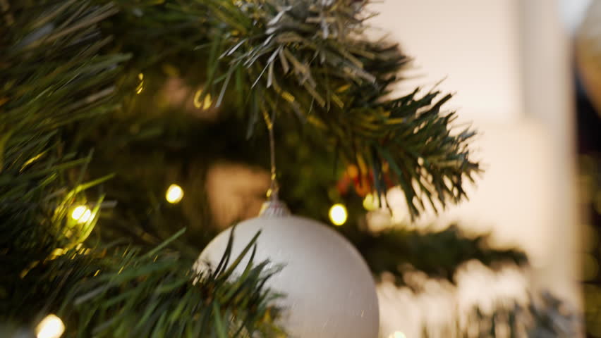 Close-up of woman hanging a white Christmas bauble on a decorated tree with warm lights. Holiday decorating at home during the festive season