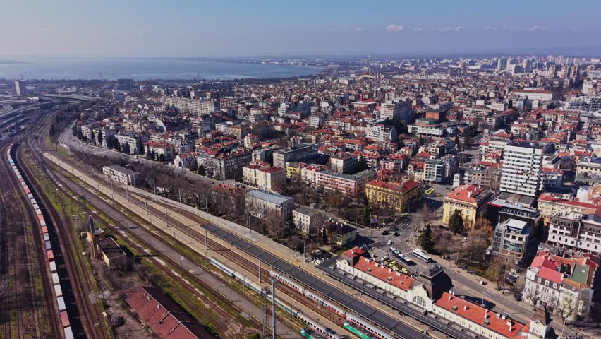High angle drone view of the urban landscape of Burgas, featuring the central railway station, tracks with trains, and red-roofed buildings under a clear sky.