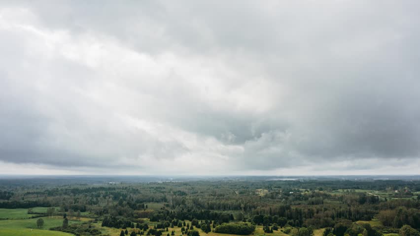 Aerial hyperlapse drone view of heavy grey storm clouds moving over vast green countryside forest and rolling fields.