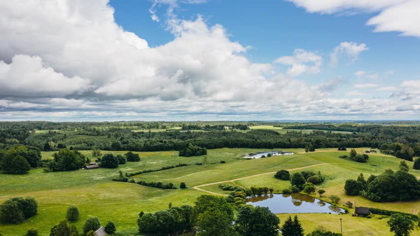 Aerial drone hyperlapse view of moving clouds over green rolling hills, fields, and ponds in scenic countryside landscape.