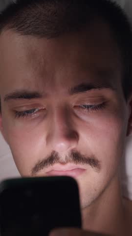 Top-down view portrait of a tired young Caucasian man lying in bed at night using his smartphone in the dark, suffering from social media addiction and insomnia.
