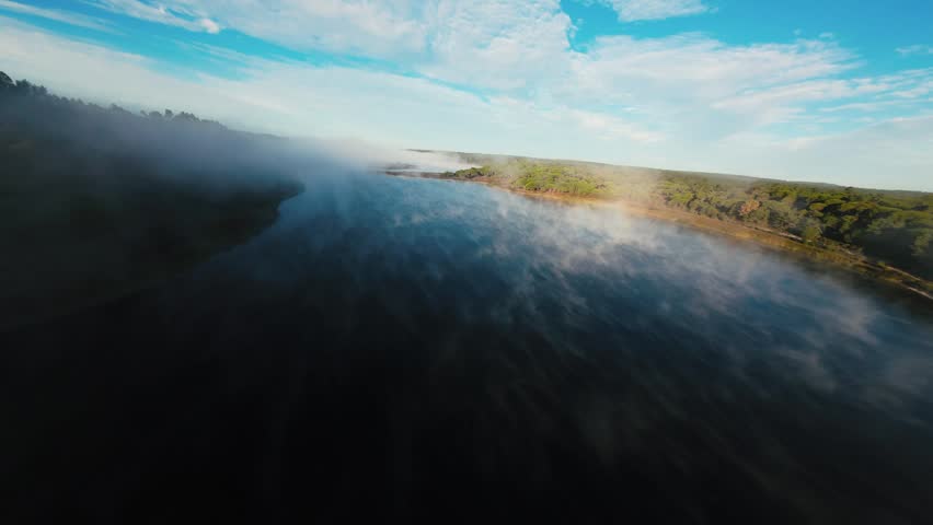 Misty river flows gently under soft dawn light. Clouds drift above green hills, framing the water