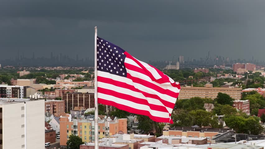 American flag waves atop pole overlooking South Orange, New Jersey rooftops near Seton Hall, brick apartment buildings and Manhattan skyline on distant stormy horizon. Aerial drone view.