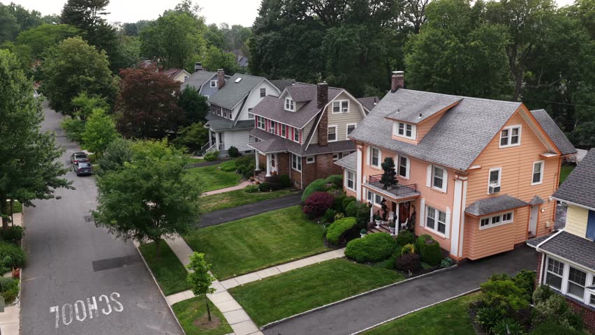 Peach colored house with balcony porch stands beside neighboring homes on tree lined street in South Orange, New Jersey, sidewalks and manicured lawns border quiet suburb. Aerial drone view.