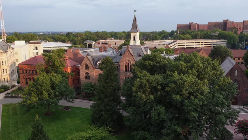 Historic brick church steeple rises above Gothic campus buildings at Seton Hall in South Orange, New Jersey, dense trees and distant dormitories surround green lawn quad. Aerial drone approaching shot