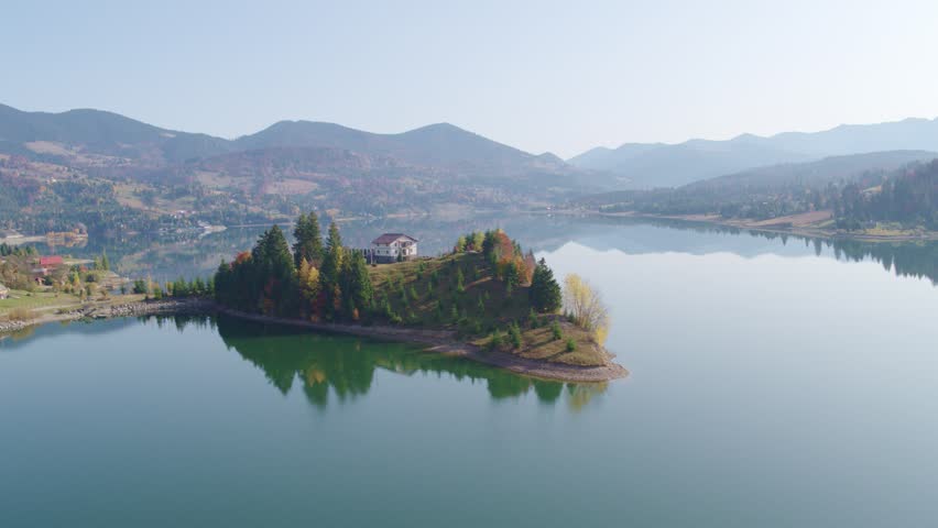 Aerial drone flight over a peninsula with a solitary house on the calm reflective waters of Colibita lake