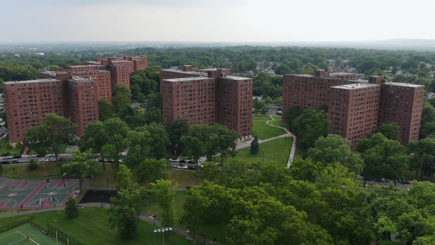 Tall brick apartment towers rise above green courtyard with basketball courts in Newark, New Jersey, tree lined paths and parked cars fill grounds near woodland edge. Aerial drone approaching shot